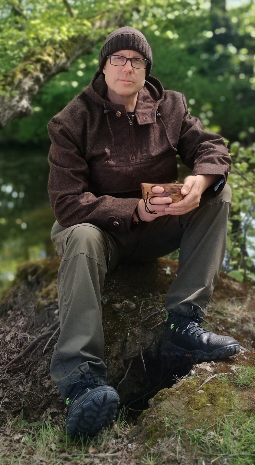 Guido Benders sitzt mit einer Holztasse im Wald auf einem Baumstamm.